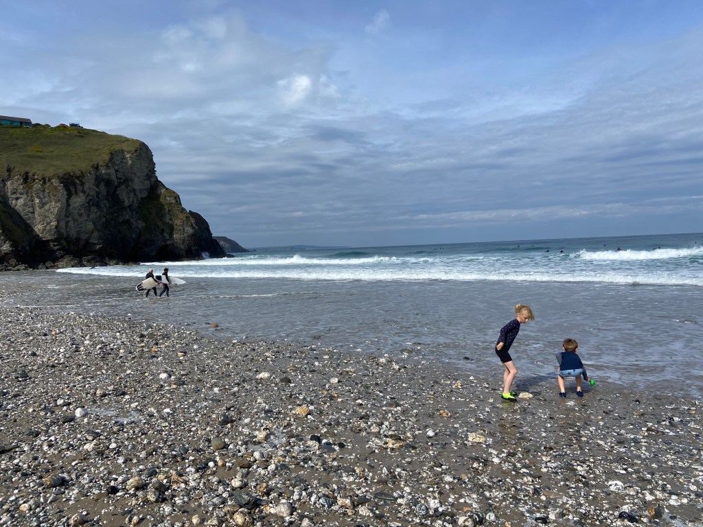 6 year old and 9 year old playing in the sea on a rocky pebble beach at Porthtowan beach in Cornwall with surfers in the background