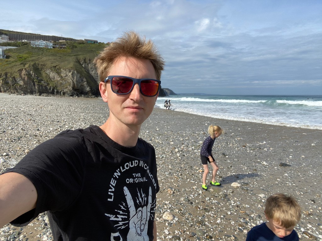 My husband in sunglasses taking a selfie at at Porthtowan beach in Cornwall with the kids playing near him
