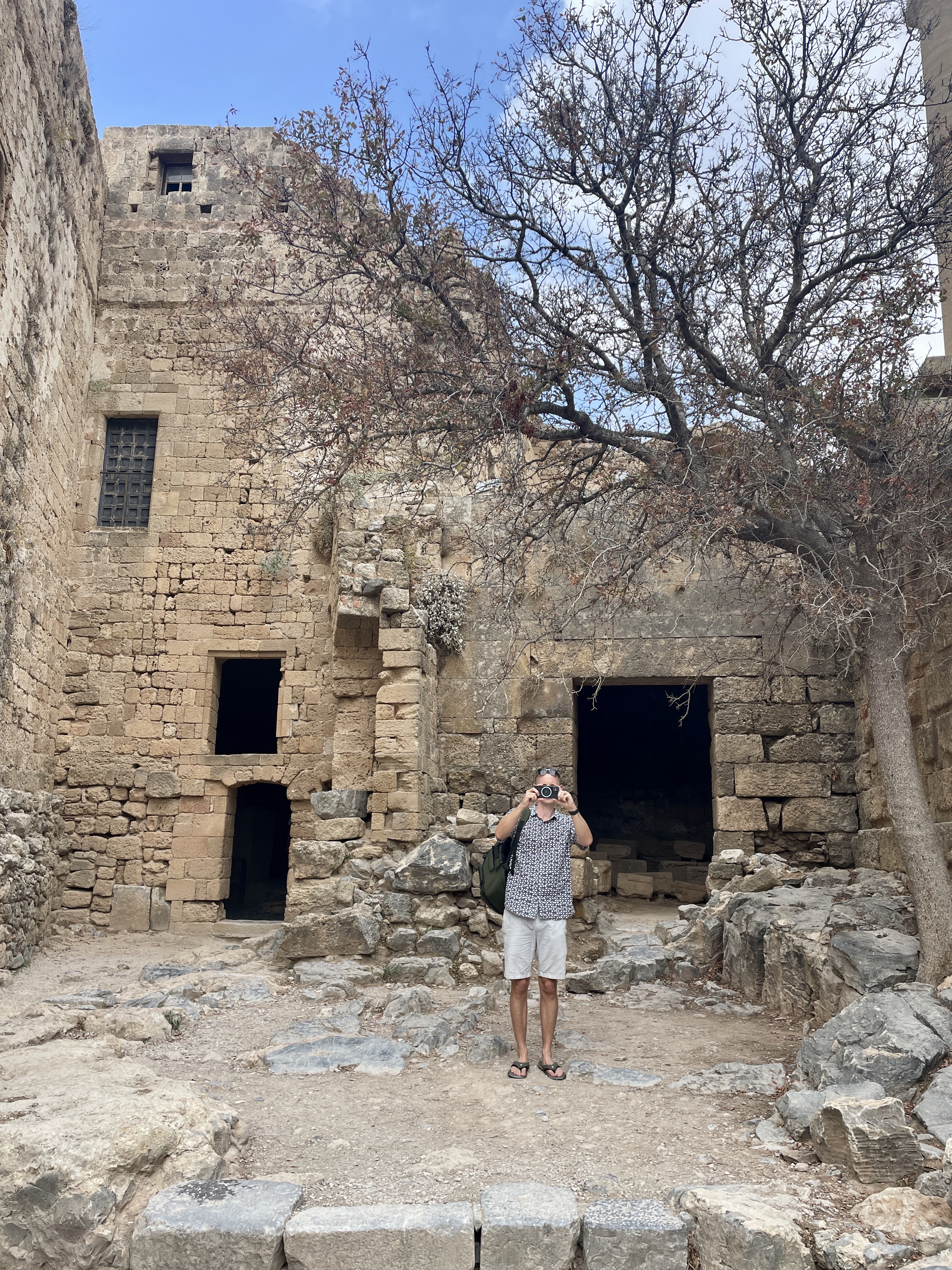 Ben taking a photo of me taking a photo of him outside some ancient walls, buildings and ruins at Lindos Acropolis